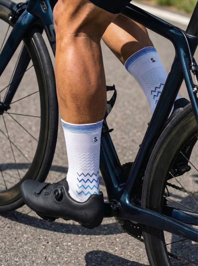 Custom branded cycling socks in white with a blue zigzag pattern, worn by a cyclist on a bike.