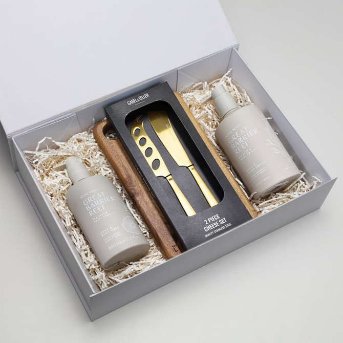 A gift box containing two cream bottles and a wooden cheese knife set, arranged on shredded paper.