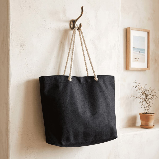 A black recycled beach tote bag with rope handles hangs on a hook against a textured wall.