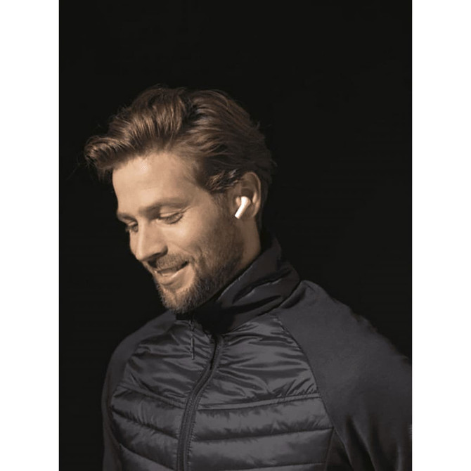 A man wearing a dark jacket smiles while using white earbuds. The background is dark, highlighting the earbuds.