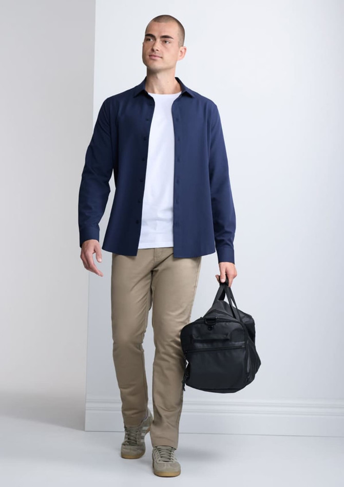 A man in a navy long sleeve shirt and beige trousers carries a black bag, standing against a neutral background.