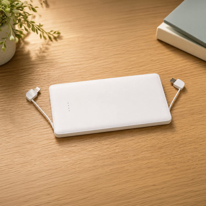 A white power bank with built-in cables on a wooden surface, accompanied by a small plant and books.