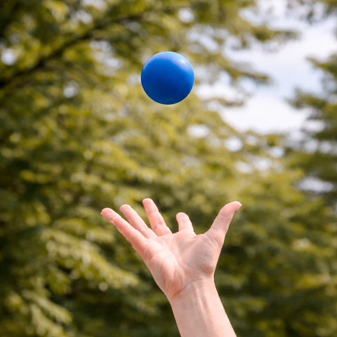 A blue Hi Bounce Ball is being tossed in the air against a blurred green background of trees.