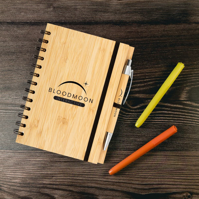 A bamboo notebook with a black elastic band, accompanied by a silver pen and colourful highlighters in orange, green, and yellow.