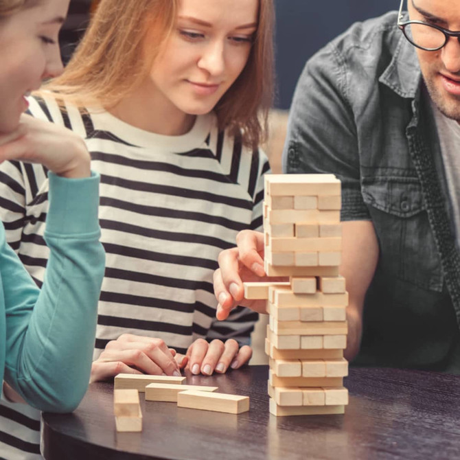 A wooden Tumbling Tower game stacked on a table, with three people engaged in play.