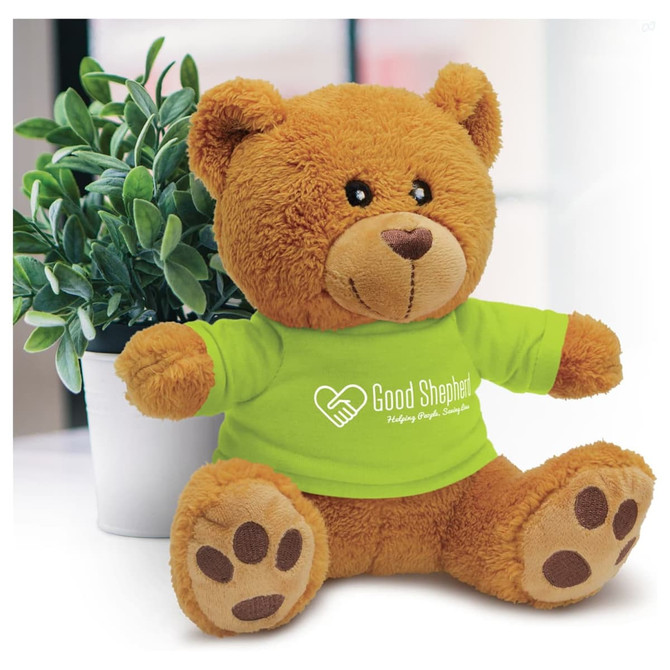 A plush teddy bear with a brown fur, wearing a green shirt and sitting next to a small potted plant.
