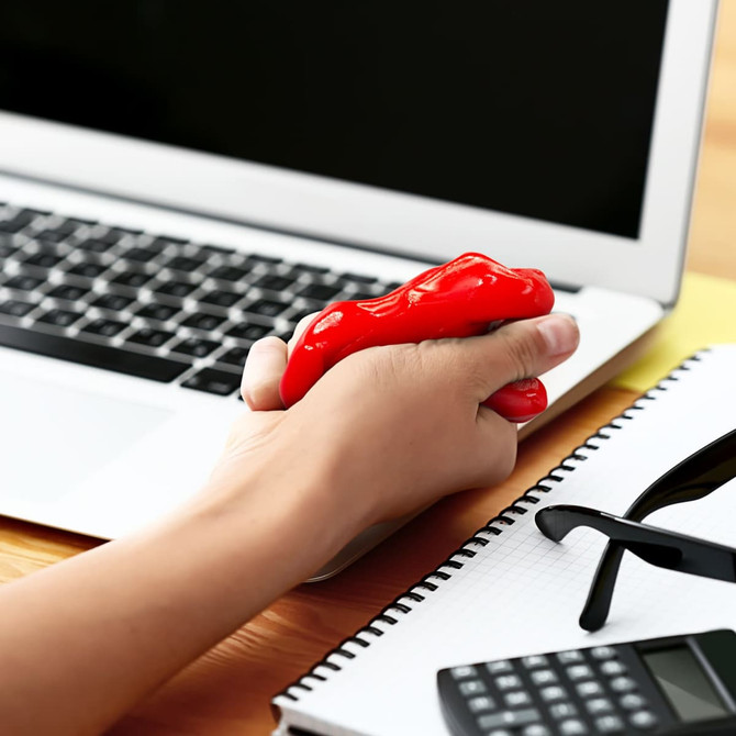 A red stress putty being squeezed in a hand, placed beside a laptop, notebook, sunglasses, and a calculator.