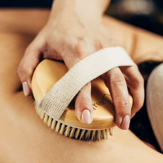 A wooden body brush with a cream strap is held against skin, featuring natural bristles for personal care. A wooden body brush with a cream strap is held against skin, featuring natural bristles for personal care.