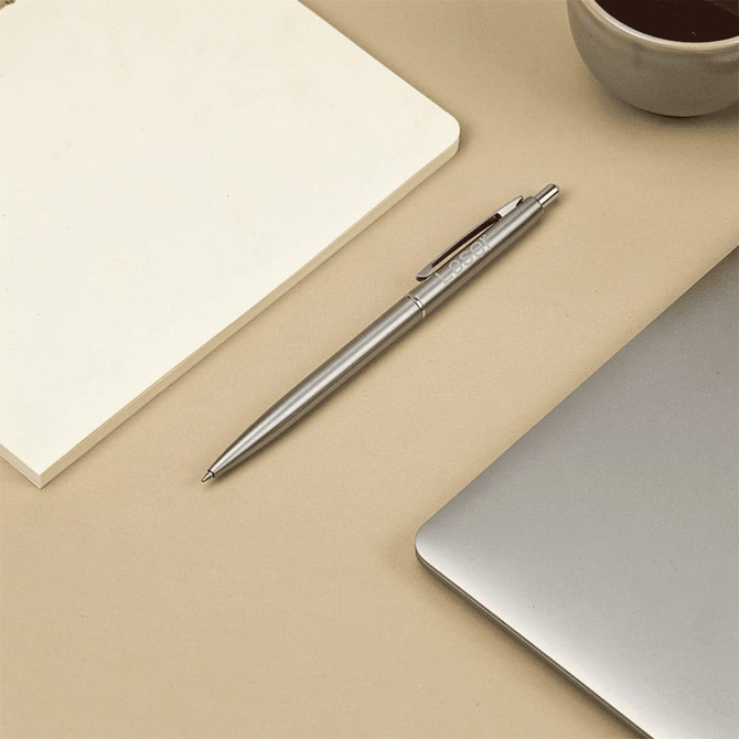 A stainless steel ballpoint pen rests beside a blank notepad and a laptop, with a cup of coffee in the background.