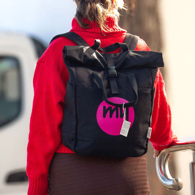 A black recycled canvas backpack tote featuring a logo, worn on the back of a person dressed in a red sweater.