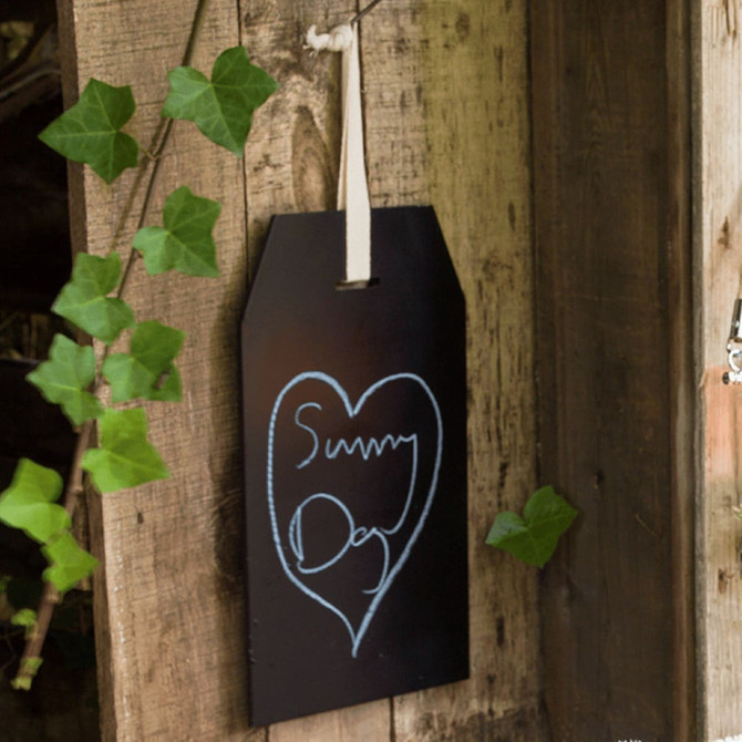A blackboard in a tag shape, suspended by a white ribbon, adorned with chalk drawings and surrounded by green ivy.