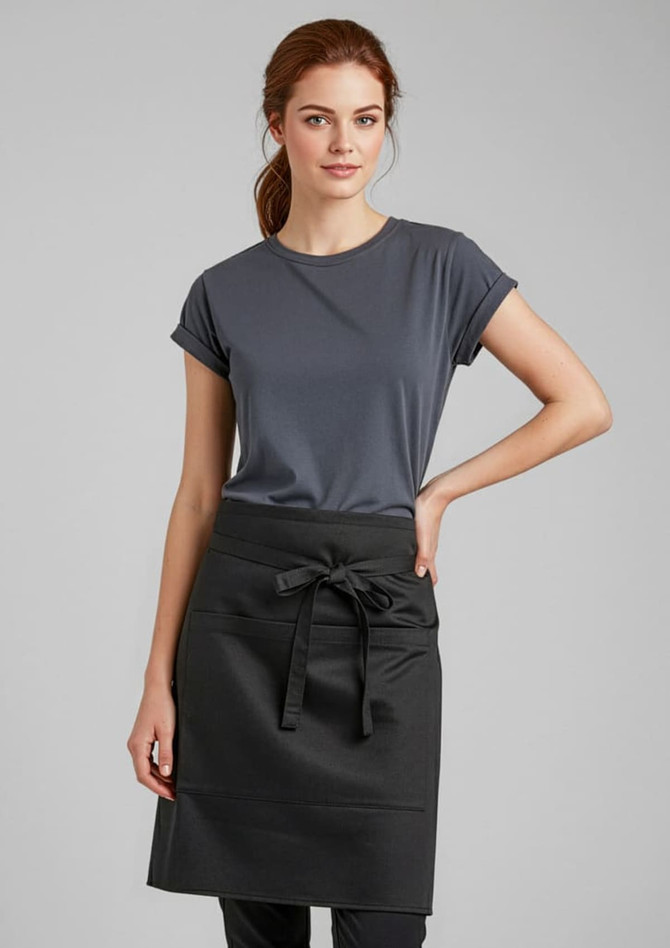 A woman in a grey t-shirt stands wearing a short waisted black apron, featuring a tied waist detail.