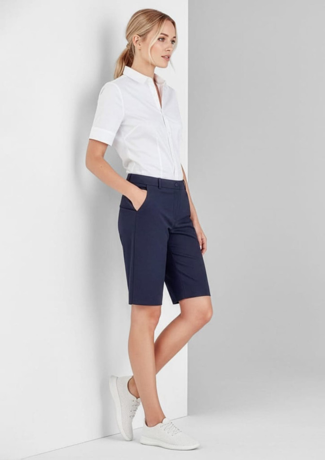 A woman wearing navy women's comfort waist cargo shorts with a white shirt and white shoes, standing against a grey wall.