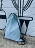 A light blue drawstring backpack with a circular pattern, featuring black cords, against a brick wall with a mural.