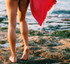 A coral red beach towel drapes over a person's legs, standing on rocky beach terrain near the water.