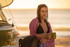 A woman in a bikini, wrapped in a purple towel, holds a green mug by a beachside van at sunset.