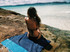 A beach towel in navy blue with a patterned edge laid on a rock, with a person sitting on it, facing the water.