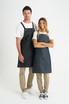 A pair of denim bib aprons in dark blue, worn by a man and a woman, featuring adjustable straps and pockets. A pair of denim bib aprons in dark blue, worn by a man and a woman, featuring adjustable straps and pockets.