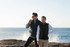 A black puffer vest worn by a man and a woman, standing by the water with a coastal background.