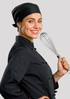 A chef in a black uniform and black bandana holds a whisk, smiling against a grey background.