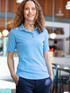 A woman stands in a café wearing a light blue polo shirt paired with dark trousers. The shirt features a logo.