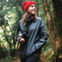 A woman wearing a dark green hooded rain jacket and a red beanie, holding a camera in a forest setting.