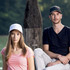 A black and a white cap are featured, worn by a man and a woman, positioned by a wooden post near water.