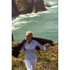A woman in a light blue button-up shirt walks along a coastal area, with cliffs and ocean visible in the background.