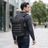 A black backpack with a logo, featuring multiple straps, worn by a man in a grey sweater, set in an urban environment.