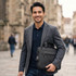 A man in a grey blazer holds a black laptop sleeve with a logo while walking in an urban setting.