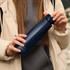 A drink bottle in navy blue, held by a person wearing a beige jacket, featuring a logo on the side. A drink bottle in navy blue, held by a person wearing a beige jacket, featuring a logo on the side.