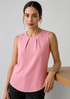 A pink sleeveless blouse with pleat detail, styled with a model standing beside a wooden surface.