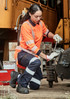 A woman in an orange high-visibility shirt and navy work pants is fixing machinery. The pants feature reflective stripes.
