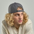 A cap made from recycled polyester in grey, worn by a model with curly blonde hair. It features a logo on the front.
