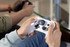 A person holding a white gaming controller with black buttons, focused on gameplay. Another controller is visible in the background.