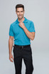 A man wearing a bright blue polo shirt and black trousers, smiling while adjusting his collar. The polo has a logo.