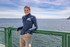 A woman stands on a ferry, wearing a blue eco fleece half zip jumper, with a scenic water view in the background. A woman stands on a ferry, wearing a blue eco fleece half zip jumper, with a scenic water view in the background.