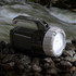A black and silver torch rests on rocky ground, with a bright light emitting from its front, surrounded by flowing water.