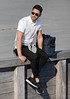 A man wearing a white polo shirt and black pants sits on a wooden railing, with a black backpack beside him.