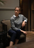 A grey knitted jumper worn over a white collared shirt, featured on a seated model in a stylish indoor setting.