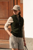 A green women's vest with a quilted pattern, worn over a beige top, paired with light grey shorts and a cap.