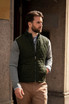 A man wearing a dark green quilted vest over a grey long-sleeve shirt, standing outside a building.