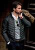 A man wearing a black quilted jacket, holding a brown bag, with sunglasses and a casual shirt underneath.