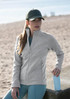 A woman wearing a light grey polar fleece jacket and a cap stands by a sandy area, holding a blue drink bottle.