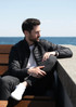 A puffer jacket in black, worn by a man seated on a bench with a seaside backdrop. The jacket features a logo.
