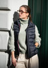 A navy vest is worn over a light green sweater, paired with cream trousers. The model has glasses and is leaning against a wall.