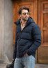 A man in a navy blue puffer jacket with a hood, wearing sunglasses and holding a bag.