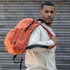 A man in a white hoodie carries an orange laptop backpack, featuring multiple compartments and a logo.