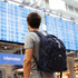 A navy blue backpack worn by a person facing an airport information board. The backpack has a logo and multiple compartments.