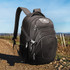 A black backpack with multiple compartments, set on a dirt path with vineyards in the background. It has a logo. A black backpack with multiple compartments, set on a dirt path with vineyards in the background. It has a logo.